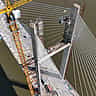 An aerial view of a yellow construction crane building a large cable-stayed bridge over a river.