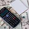 A black scientific calculator and a blank notepad sit on a scattered pile of US dollar bills.