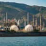 A large industrial oil refinery with spherical storage tanks and tall towers sits near a body of water.