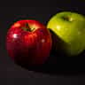 A red apple sits in sharp focus next to a blurred green apple against a dark background.
