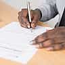 Close-up of a businessman in a grey shirt and tie signing a formal document with a fountain pen.