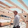 A construction worker holding a hammer stands beneath the exposed wooden roof trusses of a new home build.
