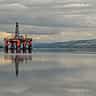 An offshore oil platform sits on calm waters beneath heavy, stormy grey clouds.