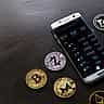 A smartphone displaying a cryptocurrency trading app surrounded by physical Bitcoin, Zcash, and other digital currency tokens on a dark surface.