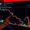 A person's finger points to a sharply falling red candlestick chart displayed on a glowing computer screen.