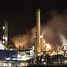 A sprawling industrial oil refinery illuminated at night, with smokestacks emitting thick plumes into the dark sky.