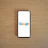 A smartphone displaying the Google logo lies flat on a light wooden surface.