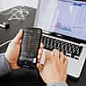 A person's hands hold a smartphone displaying a stock market application with red charts, alongside a laptop.