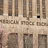 The grand stone facade of the American Stock Exchange building in New York City, partially framed by the green leaves of spring trees.