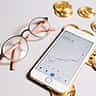 A smartphone displaying an upward-trending cryptocurrency chart lies next to gold Bitcoin tokens and stylish eyeglasses on a light surface.