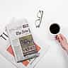 A top-down view of a black coffee cup, a folded newspaper, and reading glasses resting on a clean white desk.