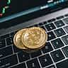 Two physical gold bitcoins rest on a laptop keyboard in front of a screen displaying a financial candlestick chart.