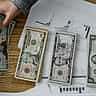 A person's hand counts US dollar bills over a desk covered in financial performance charts.