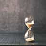 A close-up of a glass hourglass with white sand flowing downward, resting on a wooden surface.