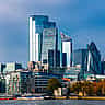 The modern financial skyline of London, featuring iconic glass skyscrapers along the River Thames under a bright blue sky.