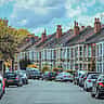 A row of classic British terraced houses stretches down a residential street beneath a partly cloudy sky.