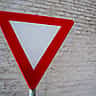 A red and white triangular yield sign stands in front of a white brick wall.