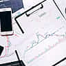Printed financial charts, a smartphone, a clipboard, and a magnifying glass sit arranged on a desk.