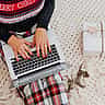 A person wearing a festive sweater and plaid pants types on a laptop next to a wrapped holiday gift on a knitted blanket.