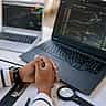 A person sits at a desk with folded hands, monitoring financial charts on two laptop screens.