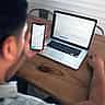 A person sits at a wooden desk analyzing financial market charts simultaneously on a smartphone and a laptop.