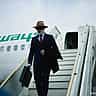 A well-dressed businessman carrying a briefcase steps down the stairs of an airplane onto the tarmac.