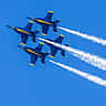 Four U.S. Navy Blue Angels F/A-18 fighter jets soar in tight formation with white smoke trails against a bright blue sky.