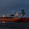 A large oil tanker ship is illuminated against a dark, moody sky at an industrial port.