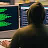 An anonymous person in a hoodie sits in a dark room working on computer monitors displaying lines of code.