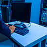 A person's hands type on a computer keyboard at a workstation inside a blue-lit server room.