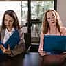 Two professional women sit side-by-side at a conference table, each reading from identical blue folders.