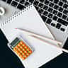 A top-down view of a modern desk featuring a laptop, grid notebook, orange calculator, and a white percent symbol.