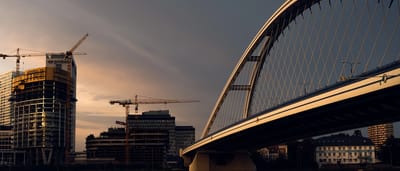 A modern arch bridge and construction cranes working on a high-rise building silhouetted against a fading sunset sky.