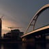 A modern arch bridge and construction cranes working on a high-rise building silhouetted against a fading sunset sky.