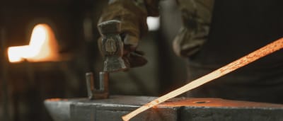A blacksmith brings a hammer down onto a glowing hot piece of metal on an anvil in a dimly lit forge.