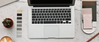 A clean, modern workspace viewed from above featuring a silver laptop, coffee, and color swatches on a white marble surface.