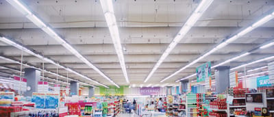 A wide, brightly lit interior of a large modern supermarket with stocked shelves and department signs.