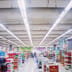 A wide, brightly lit interior of a large modern supermarket with stocked shelves and department signs.