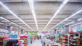 A wide, brightly lit interior of a large modern supermarket with stocked shelves and department signs.