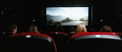 An audience sits in classic red seats watching a film on a large screen in a dark movie theater.