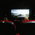 An audience sits in classic red seats watching a film on a large screen in a dark movie theater.