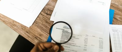 A close-up view of a hand holding a magnifying glass over printed financial documents.