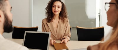 A woman in a beige blazer hands a portfolio across a table during a job interview in a bright, modern office.