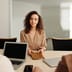 A woman in a beige blazer hands a portfolio across a table during a job interview in a bright, modern office.