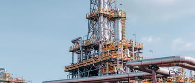 A clean, modern oil and gas refinery complex with intricate silver pipelines and tall towers against a clear blue sky.