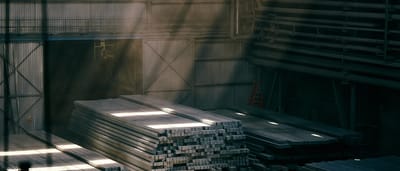 Sunlight streams into a darkened industrial warehouse, illuminating large stacks of raw steel beams.