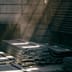 Sunlight streams into a darkened industrial warehouse, illuminating large stacks of raw steel beams.