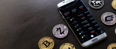A smartphone displaying a cryptocurrency trading app surrounded by physical Bitcoin, Zcash, and other digital currency tokens on a dark surface.
