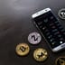 A smartphone displaying a cryptocurrency trading app surrounded by physical Bitcoin, Zcash, and other digital currency tokens on a dark surface.