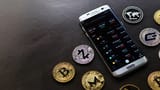 A smartphone displaying a cryptocurrency trading app surrounded by physical Bitcoin, Zcash, and other digital currency tokens on a dark surface.